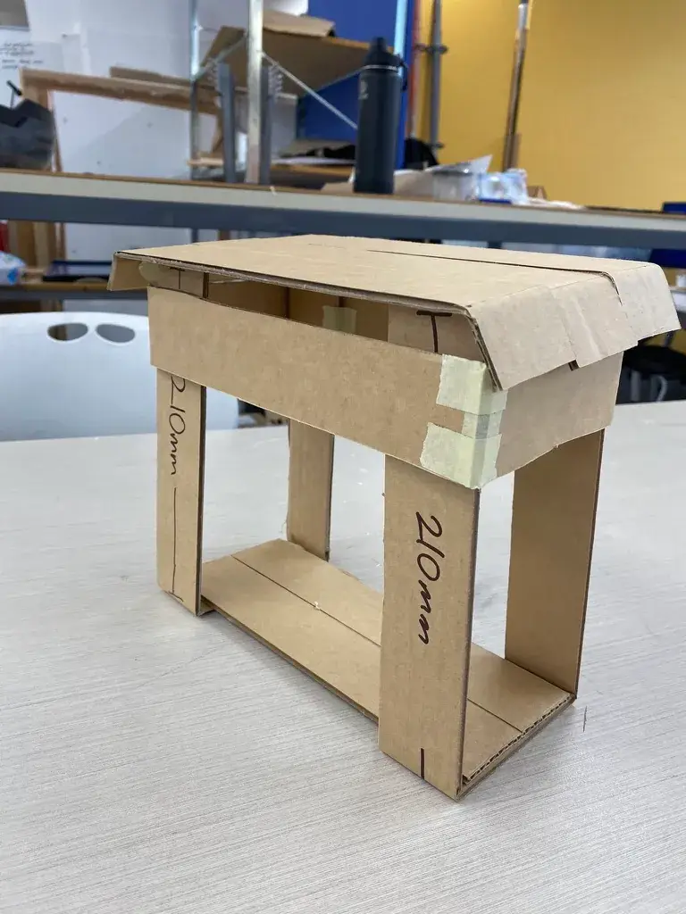 A cardboard prototype of the 'Scrappy' composting bin, showing joinery details on a workbench.