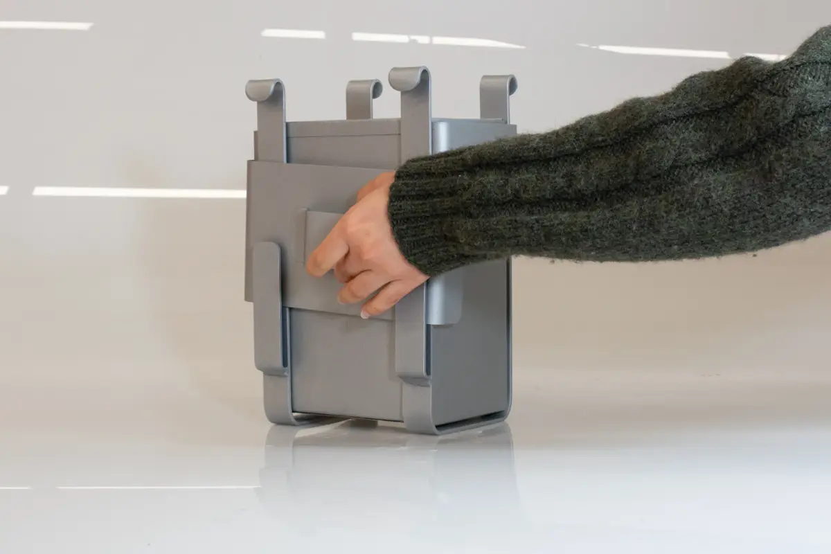 A person interacts with the fabricated metal 'Scrappy' composting bin prototype on a white surface.
