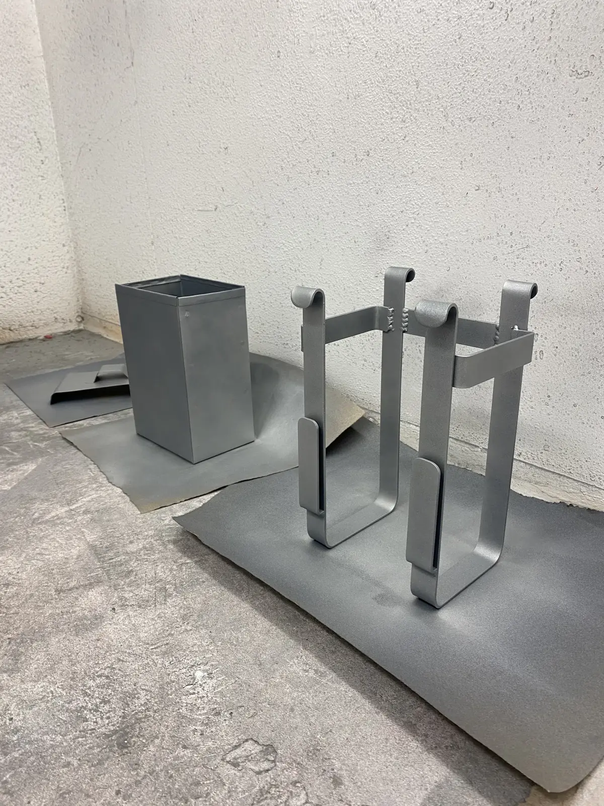 Two metal composting unit prototypes and a square container are displayed on a concrete floor.